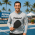 Man wearing a hoodie with 'Pickleball' text and holding a paddle on a pickleball court with palm trees in the background.